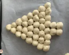 Step <p>Dispoñemos as bolas de masa nunha bandexa de forno cuberta con papel vexetal.</p><p>Colocámolas por fileiras ata conseguir a forma da árbore de Nadal.</p> illustration